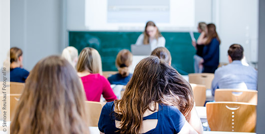 Blick von hinten über die Sitzreihen eines Seminarraums voller Studierender. Doktor Monika Voges leitet das Seminar und steht vorne neben zwei Studentinnen an der Tafel.