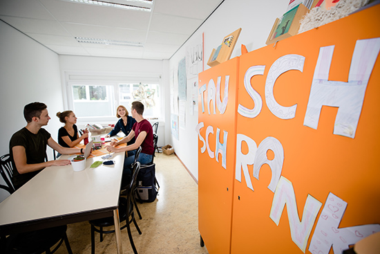 Vier Studierende sitzen an einem Tisch. Auf einen Schrank neben ihnen wurden große Buchstaben geklebt. Dort steht "Tausch Schrank".